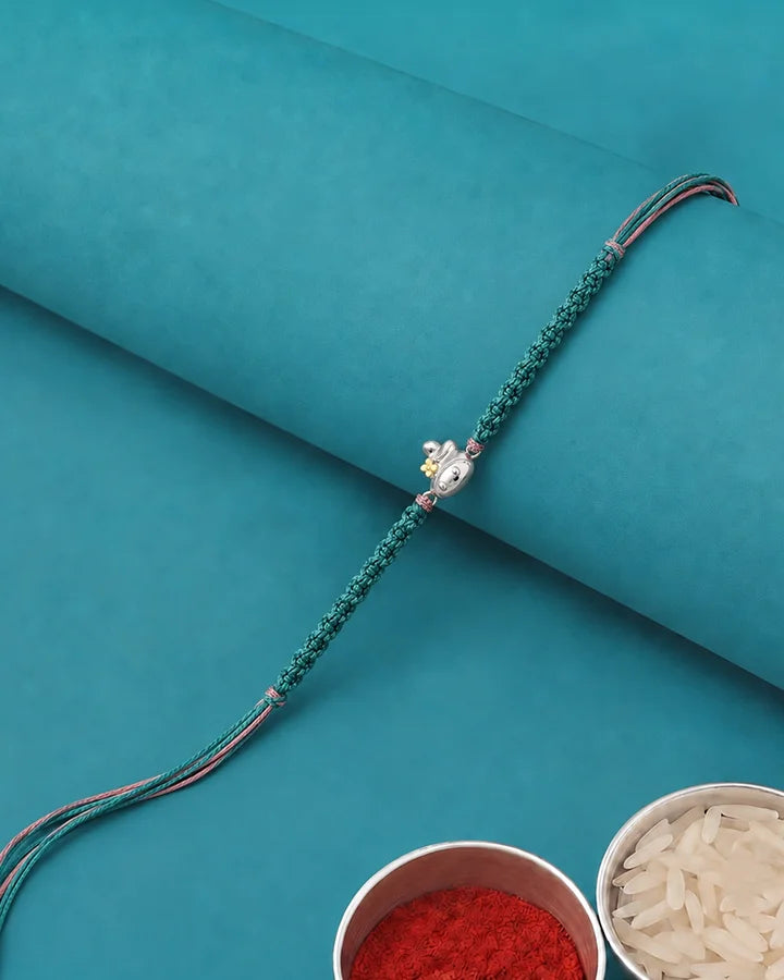 A green braided string rakhi with a small silver-toned decorative charm, laid diagonally on a teal background with two bowls at the bottom containing red powder and white grains.
