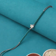 A green braided string rakhi with a small silver-toned decorative charm, laid diagonally on a teal background with two bowls at the bottom containing red powder and white grains.