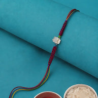 A traditional Indian Rakhi bracelet with a silver-toned Hamsa hand charm, displayed on a rolled teal paper background with bowls of red powder and rice in the foreground.
