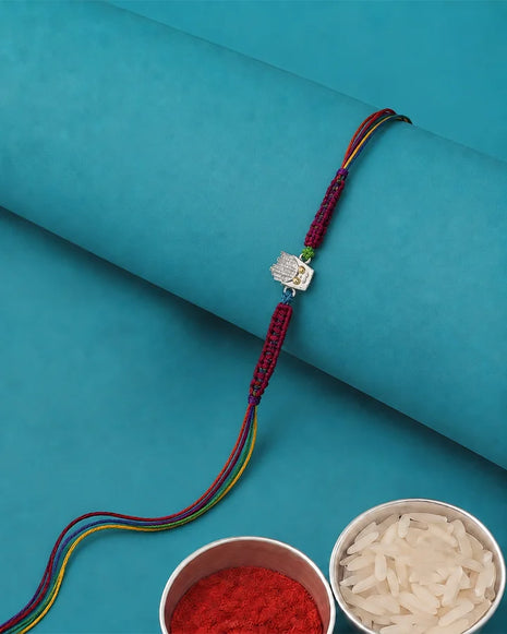 A traditional Indian Rakhi bracelet with a silver-toned Hamsa hand charm, displayed on a rolled teal paper background with bowls of red powder and rice in the foreground.