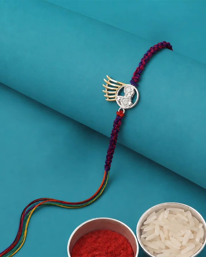 A braided Rakhi bracelet with a silver winged motif and clear stones, displayed on a teal background with bowls of red powder and white rice as props.