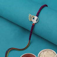 A braided Rakhi bracelet with a silver winged motif and clear stones, displayed on a teal background with bowls of red powder and white rice as props.